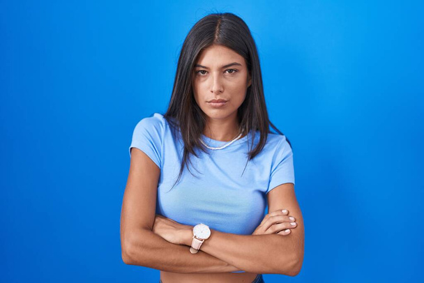 Brunette jonge vrouw staat over blauwe achtergrond sceptisch en nerveus, afkeurende uitdrukking op gezicht met gekruiste armen. negatieve persoon.  - Foto, afbeelding