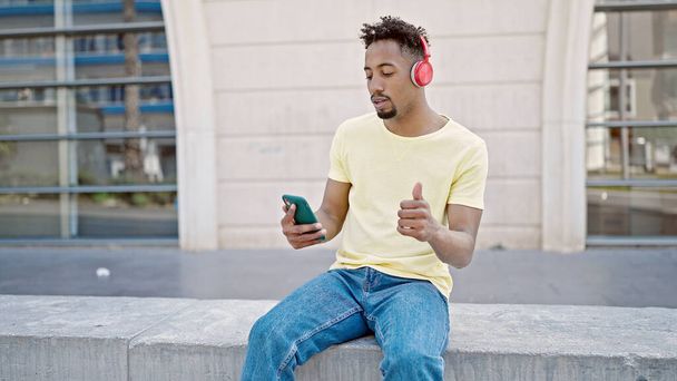 African american man listening to music using smartphone with thumb up at street - Foto, Bild