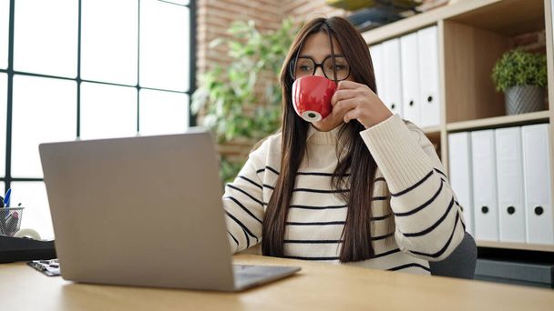 Молода красива іспанська жінка бізнесмен використовує ноутбук п'є каву в офісі
 - Фото, зображення