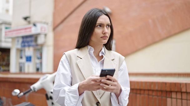 Mujer hispana hermosa joven usando teléfono inteligente con expresión seria en la calle - Foto, Imagen