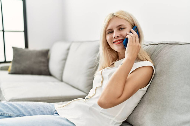 Νεαρή ξανθιά γυναίκα που μιλάει στο τηλέφωνο κάθεται στον καναπέ στο σπίτι - Φωτογραφία, εικόνα