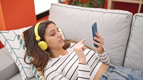 Junge schöne hispanische Frau hört zu Hause auf dem Sofa liegend Musik - Foto, Bild