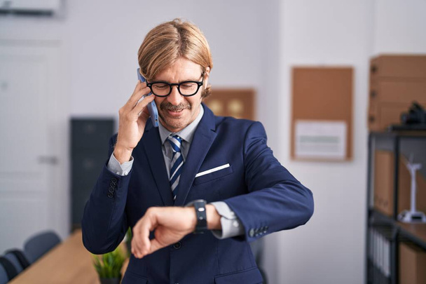 Young blond man business worker talking on smartphone looking watch at office - Фото, зображення