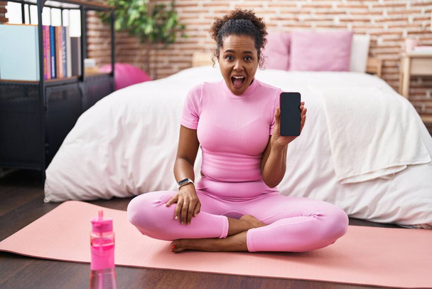 Young hispanic woman with curly hair sitting on yoga mat at the bedroom scared and amazed with open mouth for surprise, disbelief face  - Zdjęcie, obraz