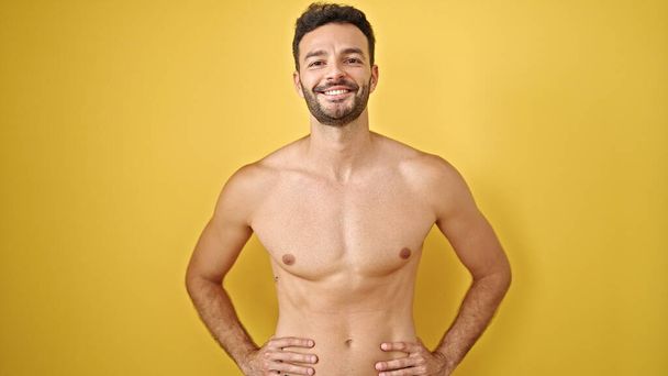 Jonge Spaanse man toerist glimlachend zelfverzekerd staand shirtloos over geïsoleerde gele achtergrond - Foto, afbeelding