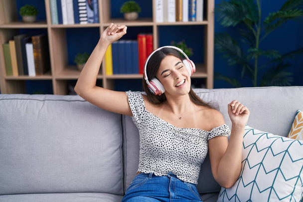 Young beautiful hispanic woman dancing and listening to music sitting on sofa at home - Photo, Image