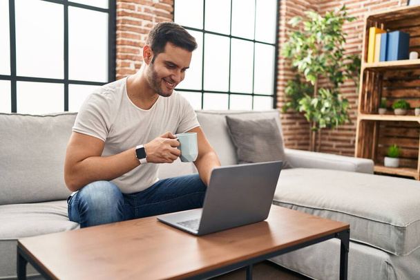 Young hispanic man using laptop and drinking coffee sitting on sofa at home - Foto, Bild