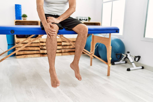 Paciente hombre de pelo gris de mediana edad que sufre de lesión de rodilla sentado en la mesa de masaje en la clínica de rehabilitación - Foto, Imagen