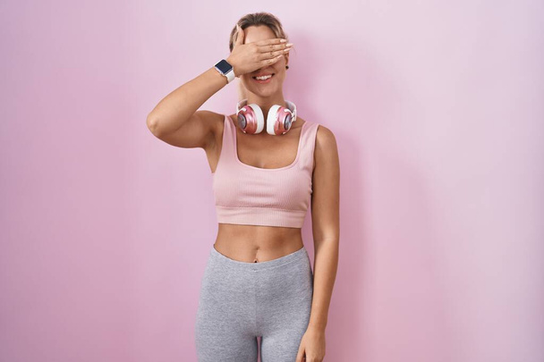 Young blonde woman wearing sportswear and headphones smiling and laughing with hand on face covering eyes for surprise. blind concept.  - Фото, изображение