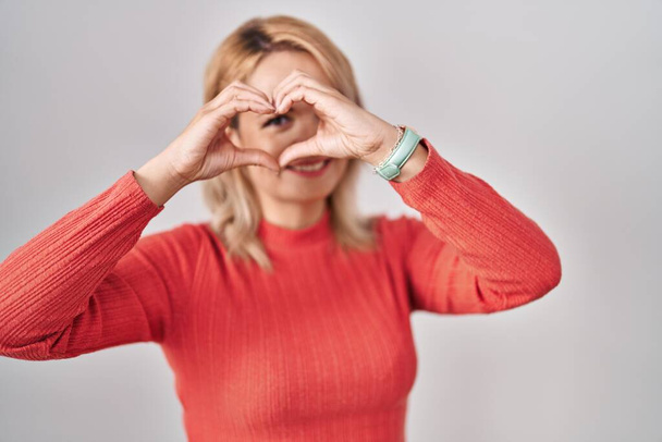 Blondýny žena stojící nad izolovaným pozadím dělá srdce tvar s rukou a prsty se usmívá při pohledu přes znamení  - Fotografie, Obrázek