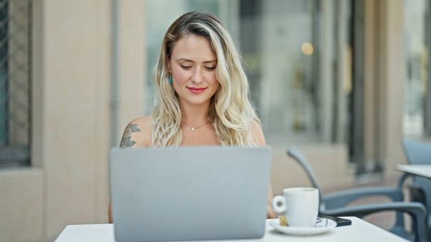 Fiatal szőke nő laptoppal ül az asztalon a kávézó teraszán - Fotó, kép