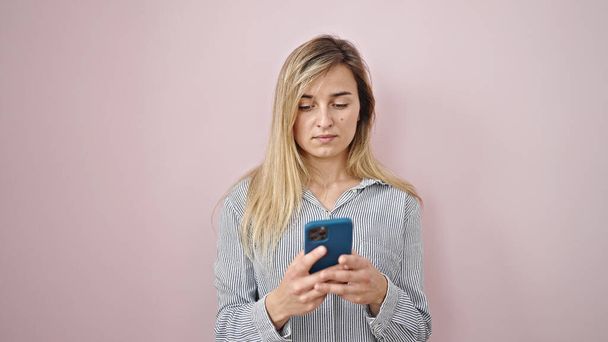 Young blonde woman using smartphone with serious expression over isolated pink background - Photo, image