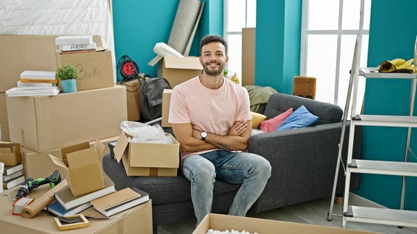 Young hispanic man smiling confident sitting on sofa with arms crossed gesture at new home - 写真・画像