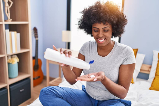 African american woman pouring lotion on hand sitting on bed at bedroom - Photo, image