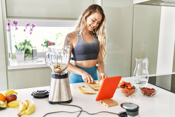 Young woman looking online juice recipe cutting banana at kitchen - Foto, afbeelding