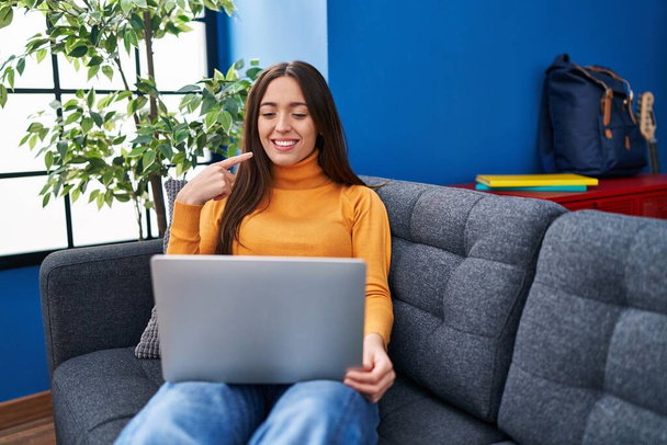 Fiatal barna nő dolgozik számítógép laptop ül a kanapén mosolyogva boldog rámutatva a kéz és az ujj  - Fotó, kép