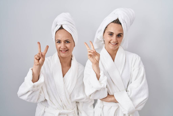 Middle age woman and daughter wearing white bathrobe and towel smiling with happy face winking at the camera doing victory sign. number two.  - Fotoğraf, Görsel