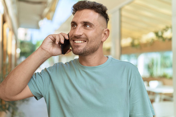 Νεαρός Ισπανόφωνος άντρας χαμογελάει με αυτοπεποίθηση μιλώντας στο smartphone στη βεράντα της καφετέριας - Φωτογραφία, εικόνα