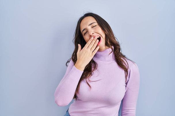 Young brunette woman standing over blue background bored yawning tired covering mouth with hand. restless and sleepiness.  - Foto, Bild