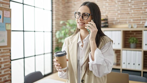 Молодая красивая латиноамериканка бизнес-работница разговаривает по смартфону, пьет кофе в офисе - Фото, изображение