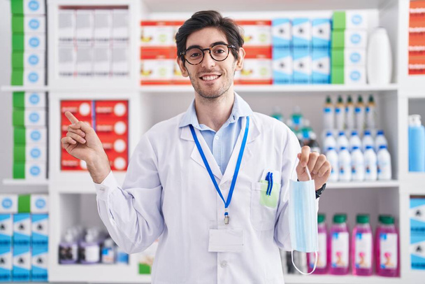 Fiatal spanyol férfi dolgozik gyógyszertár gyógyszertár biztonsági maszk mosolyogva boldog mutató kéz és ujj az oldalon  - Fotó, kép