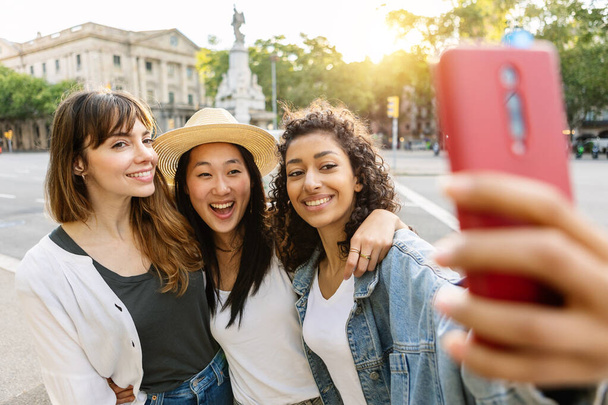 Young diverse female friends enjoying summer days taking selfie together outdoors. Three beautiful multiracial girls laughing while having fun on travel vacation in Barcelona. Friendship concept. - Фото, изображение
