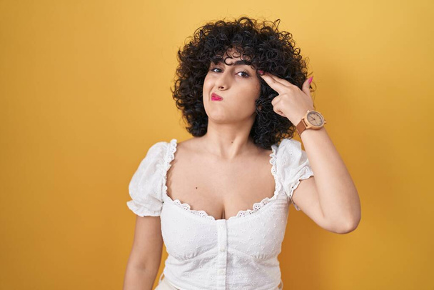 Young brunette woman with curly hair standing over yellow background shooting and killing oneself pointing hand and fingers to head like gun, suicide gesture.  - Photo, Image