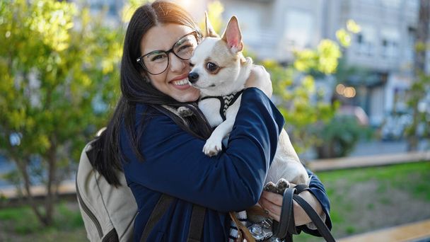 Young hispanic woman with chihuahua dog wearing backpack smiling together at park - Photo, Image