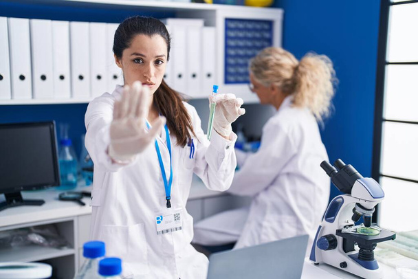 Young hispanic woman working at scientist laboratory with open hand doing stop sign with serious and confident expression, defense gesture  - Foto, Imagem