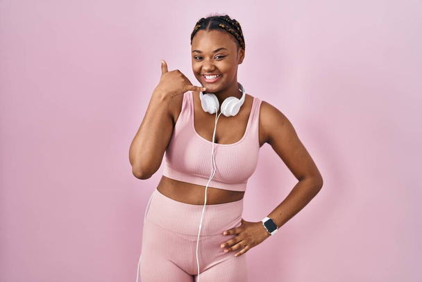 Afroamerykanka w warkoczach, ubrana w sportową odzież i ze słuchawkami, uśmiechająca się, robiąca telefoniczne gesty ręką i palcami, jakby rozmawiała przez telefon. komunikowanie pojęć.  - Zdjęcie, obraz