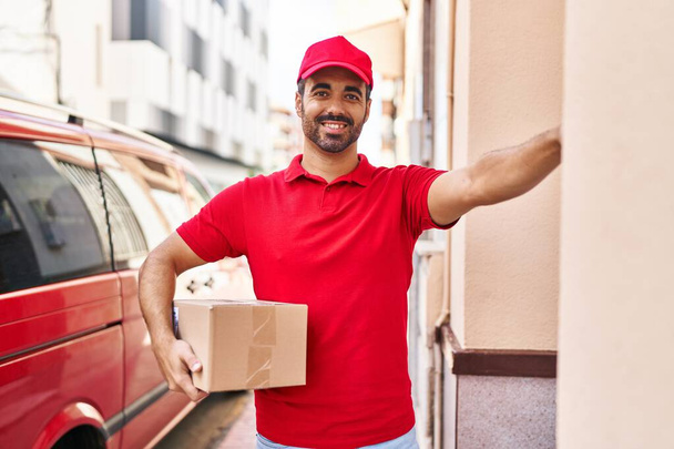 Young hispanic man courier holding package press doorbell at street - Fotó, kép