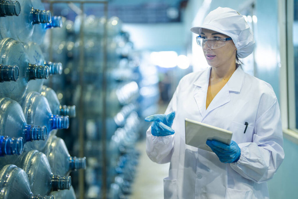 Dělník z továrny kontroluje láhve s vodou ve skladu v průmyslové továrně. Pracovnice zaznamenávající údaje na výrobní lince nápojů. - Fotografie, Obrázek