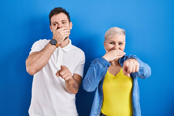 Junge brasilianische Mutter und ihr Sohn stehen vor blauem Hintergrund und lachen dich an, zeigen mit der Hand über den Mund in die Kamera, schämen sich.  - Foto, Bild