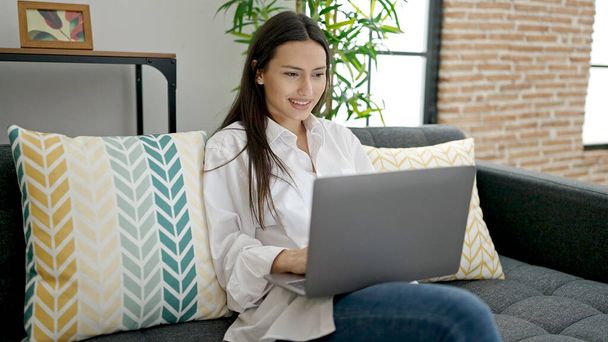 Nuori kaunis latino nainen käyttää kannettavaa tietokonetta istuu sohvalla kotona - Valokuva, kuva