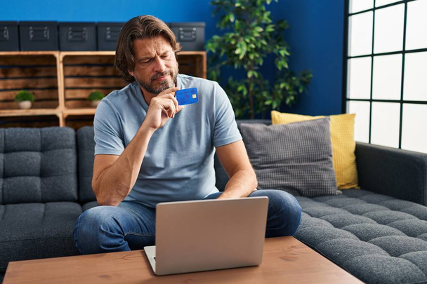 Uomo di mezza età utilizzando laptop e carta di credito con espressione dubbio a casa - Foto, immagini