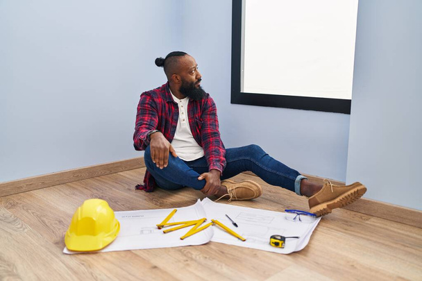 Africký Američan sedí na podlaze v novém domově a dívá se na nákresy rozhlížející se na stranu, relaxační profil pózuje s přirozenou tváří s sebevědomým úsměvem.  - Fotografie, Obrázek