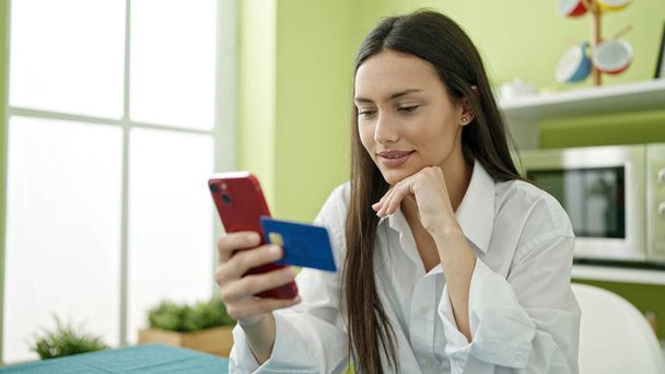 Młoda piękna Latynoska kobieta zakupy ze smartfonem i kartą kredytową siedzi na stole w jadalni - Zdjęcie, obraz