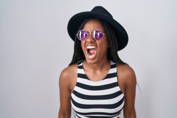 Jovem afro-americana com tranças usando chapéu e óculos de sol com raiva e gritos loucos frustrados e furiosos, gritando com raiva. raiva e conceito agressivo.  - Foto, Imagem