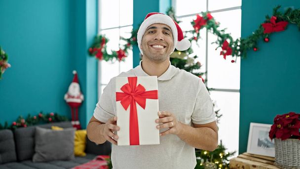 Junger hispanischer Mann lächelt zuversichtlich und hält Weihnachtsgeschenk zu Hause - Foto, Bild