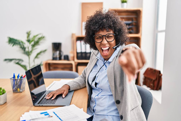 Zwarte vrouw met krullend haar in callcenter agent headset op kantoor wijzend ontevreden en gefrustreerd naar de camera, boos en woedend op jou  - Foto, afbeelding