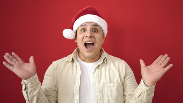 Νεαρός ισπανόφωνος άνδρας έκπληξη έκφραση φορώντας χριστουγεννιάτικο καπέλο πάνω από απομονωμένο κόκκινο φόντο - Φωτογραφία, εικόνα
