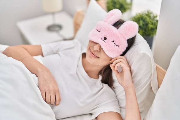 Giovane donna ispanica indossa divertente maschera del sonno sdraiato sul letto dormire in camera da letto - Foto, immagini