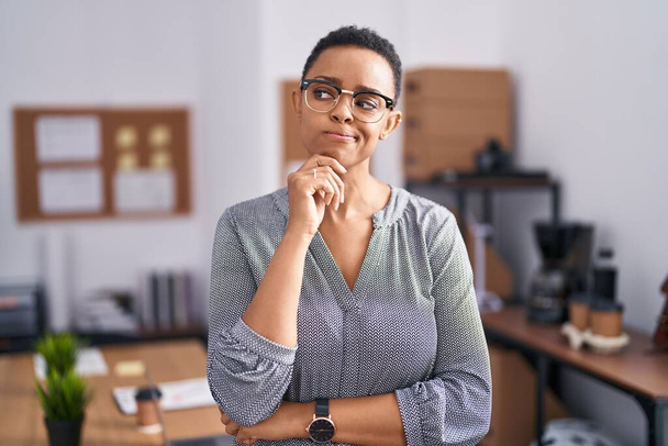 オフィスで働くアフリカ系アメリカ人の女性は、顎の質問について考える上で手で眼鏡をかけて、集中的な表情。思慮深い顔で笑顔。疑わしい概念.  - 写真・画像