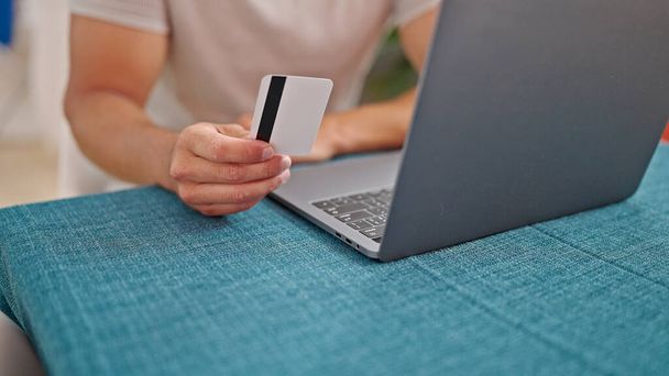 Молодой латиноамериканец, использующий ноутбук с кредитной картой, сидит за столом в столовой - Фото, изображение