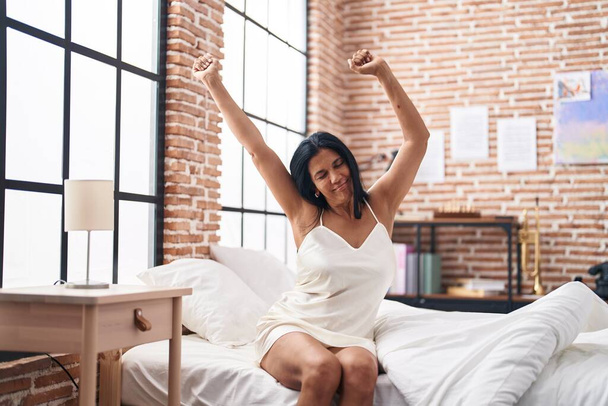 Middelbare leeftijd Spaanse vrouw wakker stretching armen in de slaapkamer - Foto, afbeelding