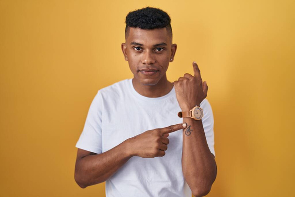 Young hispanic man standing over yellow background in hurry pointing to watch time, impatience, looking at the camera with relaxed expression  - Photo, Image
