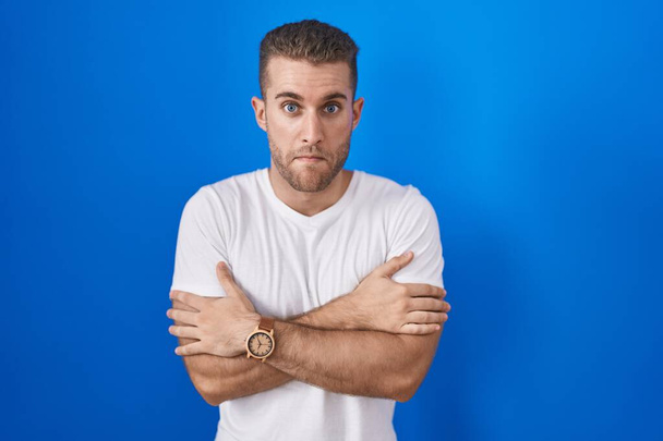 Young caucasian man standing over blue background shaking and freezing for winter cold with sad and shock expression on face  - Фото, изображение