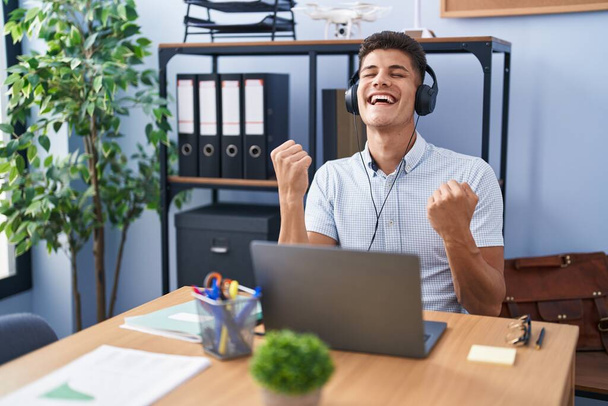 Young hispanic man working at the office wearing headphones very happy and excited doing winner gesture with arms raised, smiling and screaming for success. celebration concept.  - Photo, Image
