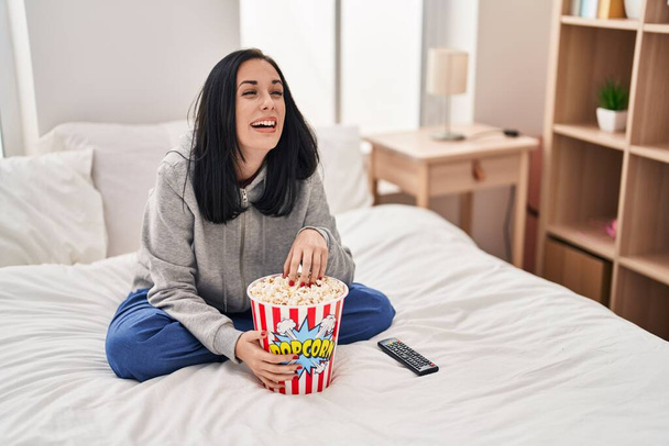 Hispanic woman eating popcorn watching a movie on the bed smiling and laughing hard out loud because funny crazy joke.  - 写真・画像