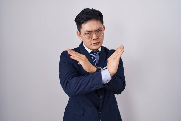 Молодий азіатський чоловік у діловому костюмі та вираженні відторгнення краватки перетинає руки, роблячи негативний знак, сердите обличчя  - Фото, зображення